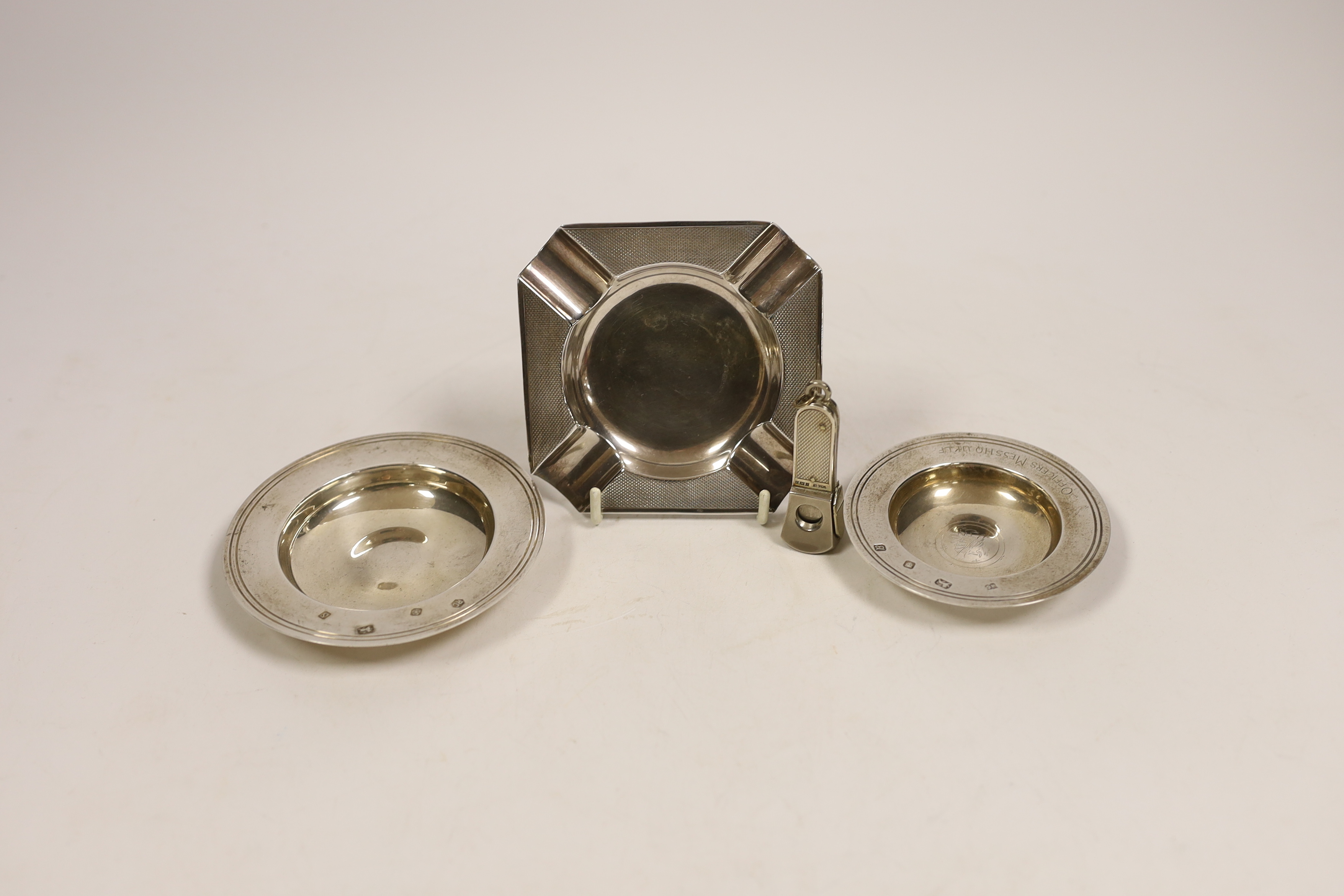 Three assorted silver ashtrays, including one engraved 'Officers Mess HQ UKLF', largest 10.1cm, together with a silver mounted cigar cutter.